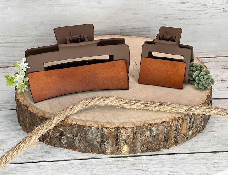 May include: Two brown plastic hair clips with a brown leather-like insert. The larger clip is curved and the smaller clip is rectangular. Both clips are on a wooden surface.