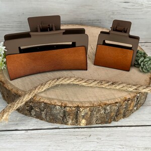 May include: Two brown plastic hair clips with a brown leather-like insert. The larger clip is curved and the smaller clip is rectangular. Both clips are on a wooden surface.