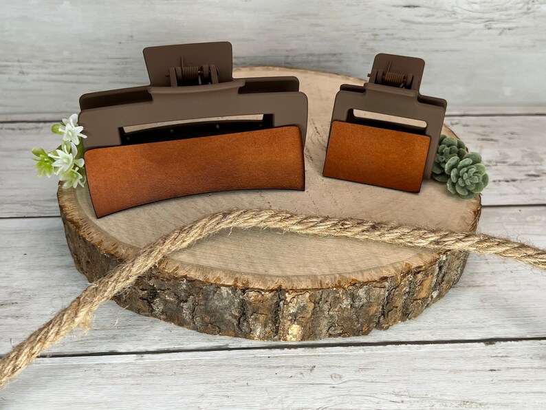 May include: Two brown plastic hair clips with a brown leather-like insert. The larger clip is curved and the smaller clip is rectangular. Both clips are on a wooden surface.