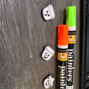 May include: Four small, white and black Halloween-themed magnets shaped like buckets with jack-o'-lantern faces. Two paint markers, one orange and one green, are also in the image. The markers have the word "painters" printed on them.