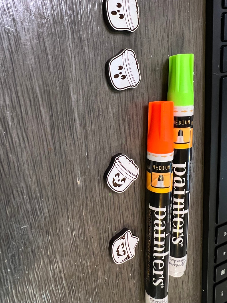 May include: Four small, white and black Halloween-themed magnets shaped like buckets with jack-o'-lantern faces. Two paint markers, one orange and one green, are also in the image. The markers have the word "painters" printed on them.