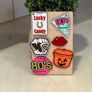 May include: A wooden board with various Halloween-themed decorations, including a horseshoe with the text "Lucky U Candy", a red lip, a vampire's mouth, a pumpkin bucket, and two signs that say "90's Halloween" and "80's Halloween".
