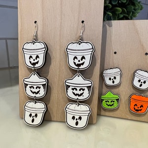 May include: Halloween-themed earrings and studs. The earrings feature a three-tiered design of smiling jack-o'-lantern buckets. Studs include two jack-o'-lantern buckets, one green, and one orange. All are black and white with a smiling face.