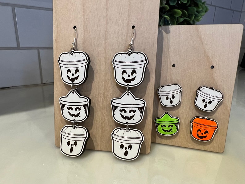 May include: Halloween-themed earrings and studs. The earrings feature a three-tiered design of smiling jack-o'-lantern buckets. Studs include two jack-o'-lantern buckets, one green, and one orange. All are black and white with a smiling face.