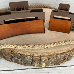 May include: Two brown hair clips with a brown suede-like covering. The larger clip is curved and the smaller clip is rectangular. The clips are on a wooden slice with a rope wrapped around it.