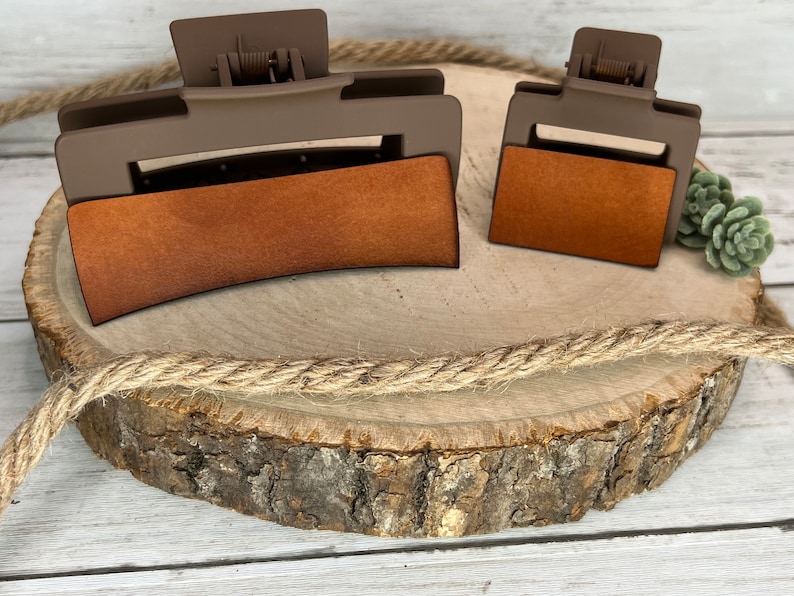 May include: Two brown hair clips with a brown suede-like covering. The larger clip is curved and the smaller clip is rectangular. The clips are on a wooden slice with a rope wrapped around it.