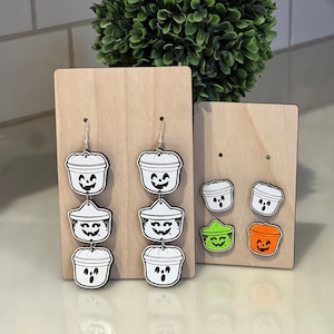 May include: Wooden Halloween-themed earrings and stud earrings. The earrings are shaped like jack-o'-lantern buckets with smiling faces. The stud earrings are smaller versions of the jack-o'-lantern buckets, with green and orange accents.