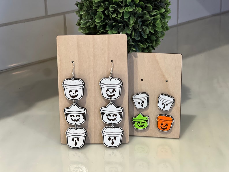 May include: Wooden Halloween-themed earrings and stud earrings. The earrings are shaped like jack-o'-lantern buckets with smiling faces. The stud earrings are smaller versions of the jack-o'-lantern buckets, with green and orange accents.