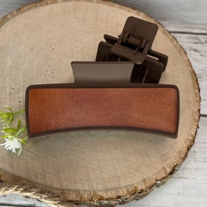 May include: A brown plastic hair claw with a textured, faux leather top. The claw is designed to hold hair in place.
