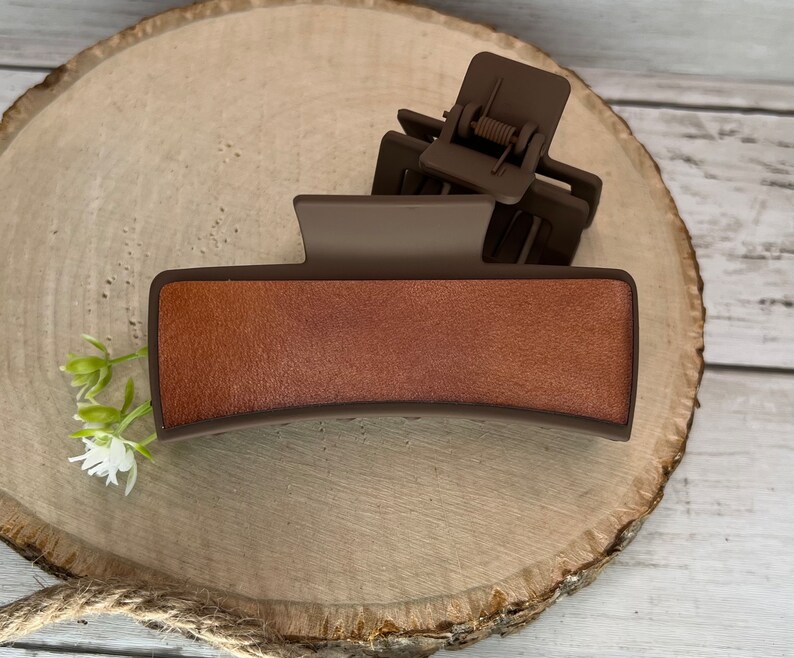 May include: A brown plastic hair claw with a textured, faux leather top. The claw is designed to hold hair in place.