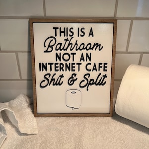 May include: A white wooden sign with a brown frame that reads "This is a Bathroom Not an Internet Cafe Shit & Split". A cartoon toilet paper roll is below the text.