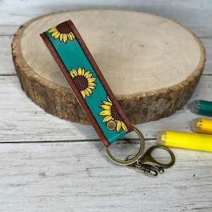 May include: A teal leather keychain with a sunflower pattern and a brown leather border. The keychain has a brass key ring and a lobster clasp.