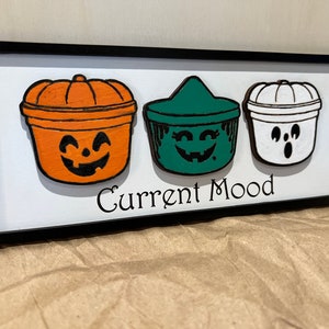 May include: A framed wooden sign with three painted pumpkin-shaped containers. The containers are painted orange, green, and white with different facial expressions. The sign reads "Current Mood".