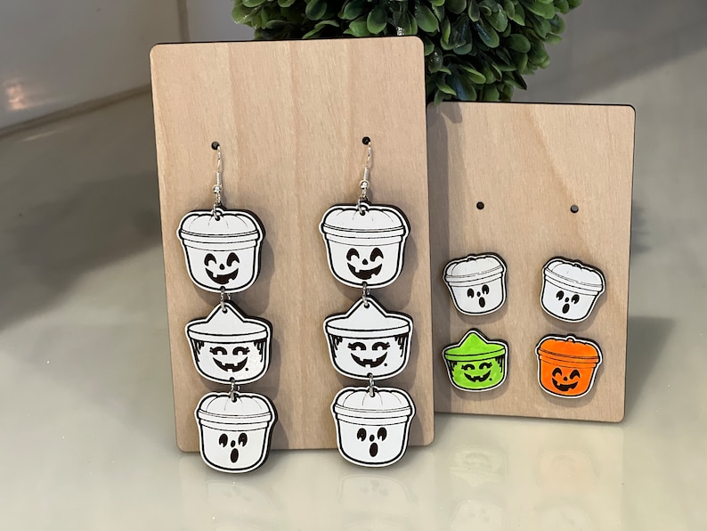 May include: Wooden display featuring Halloween-themed earrings and studs. The earrings are white with black outlines, depicting jack-o'-lantern buckets. Studs in green and orange are also displayed. The earrings are on a wooden display.