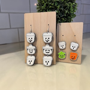 May include: Wooden Halloween-themed earrings and stud earrings. The earrings feature a stacked design of white and black jack-o'-lantern buckets. The stud earrings include green and orange jack-o'-lantern buckets. The earrings are displayed on wooden stands.