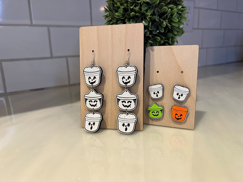 May include: Wooden Halloween-themed earrings and stud earrings. The earrings feature a stacked design of white and black jack-o'-lantern buckets. The stud earrings include green and orange jack-o'-lantern buckets. The earrings are displayed on wooden stands.