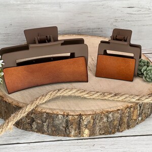 May include: Two brown plastic hair clips with brown leather-like inserts. The larger clip is curved and the smaller clip is rectangular. The clips are on a wooden surface.