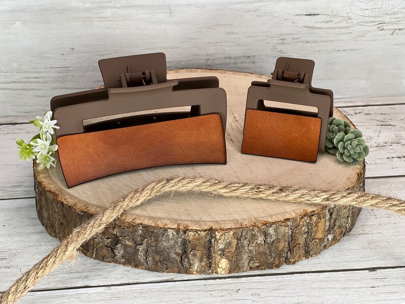May include: Two brown plastic hair clips with brown leather-like inserts. The larger clip is curved and the smaller clip is rectangular. The clips are on a wooden surface.