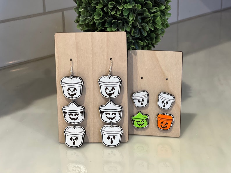 May include: Wooden display boards showcasing Halloween-themed earrings and pins. The earrings feature a cascading design of white buckets with smiling jack-o'-lantern faces. Pins include white, green, and orange jack-o'-lantern buckets.
