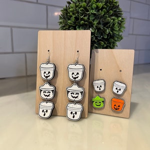 May include: Wooden Halloween-themed earrings and stud earrings. The earrings feature a white and black design of a bucket with a smiling face. The stud earrings are in the shape of a bucket with a smiling face, in green and orange.