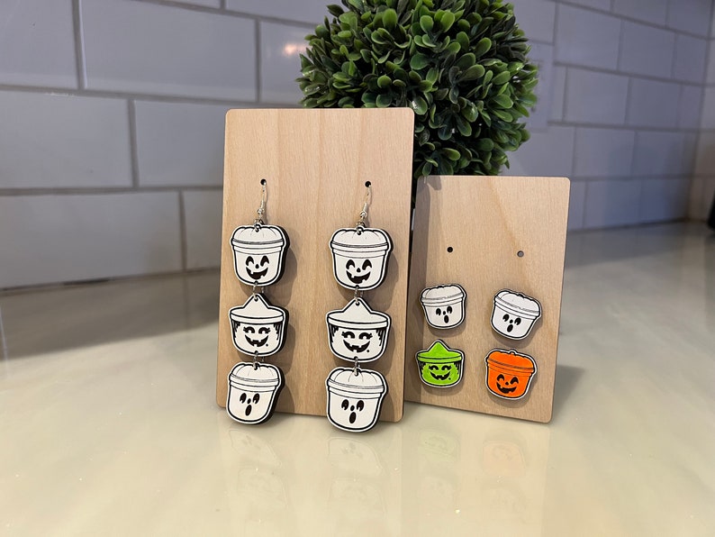 May include: Wooden Halloween-themed earrings and stud earrings. The earrings feature a white and black design of a bucket with a smiling face. The stud earrings are in the shape of a bucket with a smiling face, in green and orange.