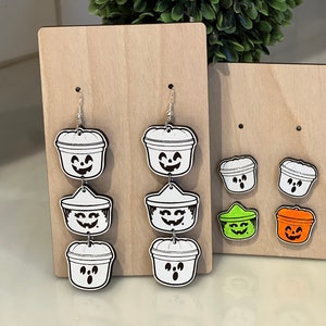 May include: Wooden display featuring Halloween-themed earrings and studs. The earrings are white with black outlines, depicting jack-o'-lantern buckets. Studs in green and orange are also displayed. The earrings are on a wooden display.