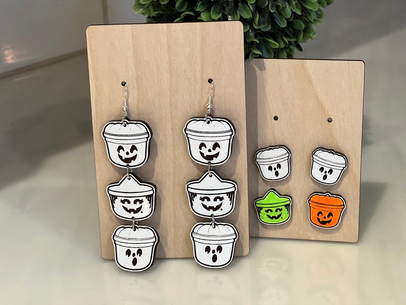 May include: Wooden display featuring Halloween-themed earrings and studs. The earrings are white with black outlines, depicting jack-o'-lantern buckets. Studs in green and orange are also displayed. The earrings are on a wooden display.