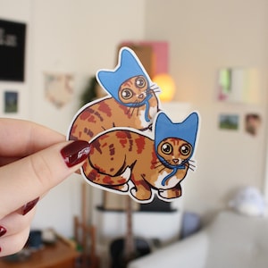 Könnte beinhalten: Zwei Katzenaufkleber. Jeder Aufkleber zeigt eine Cartoon-Katze mit orange-braunem Fell, die einen blauen Hut trägt. Die Katzen haben große Augen und sind in einem niedlichen Stil gezeichnet. Die Aufkleber werden von einer Hand gehalten.