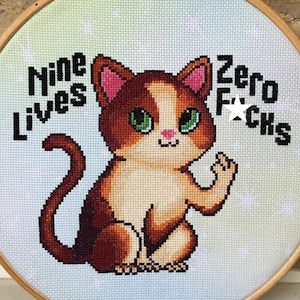 May include: A cross-stitch embroidery of a cartoon cat with green eyes and a pink nose. The cat is sitting with its right paw raised in a fist. The text "Nine Lives Zero F*cks" is stitched above the cat. The embroidery is framed in a wooden hoop.