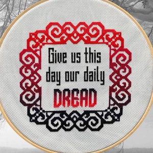 May include: A cross-stitch embroidery hoop with the text "Give us this day our daily DREAD" in red and black. The design is framed by a decorative border. The background is a misty, grayscale forest scene.