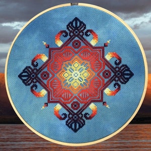 Puede incluir: Un diseño de mandala cosido a mano en tonos de rojo, naranja, amarillo y negro, enmarcado en un aro azul. El diseño está cosido en tela Aida azul teñida a mano de 18ct. "Ejemplo cosido [Aida azul teñido a mano 18ct]" está escrito debajo del aro.