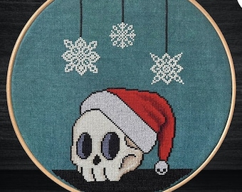 May include: Cross stitch pattern for a skull wearing a Santa hat with snowflakes hanging above it. The pattern is stitched on teal fabric in an 8 inch hoop.