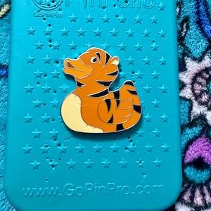 May include: A pin featuring a cartoon tiger duck design, with orange and black stripes, set against a teal background with star-shaped cutouts. The pin is on a teal PinFolio case. The website www.GoPinPro.com is visible.