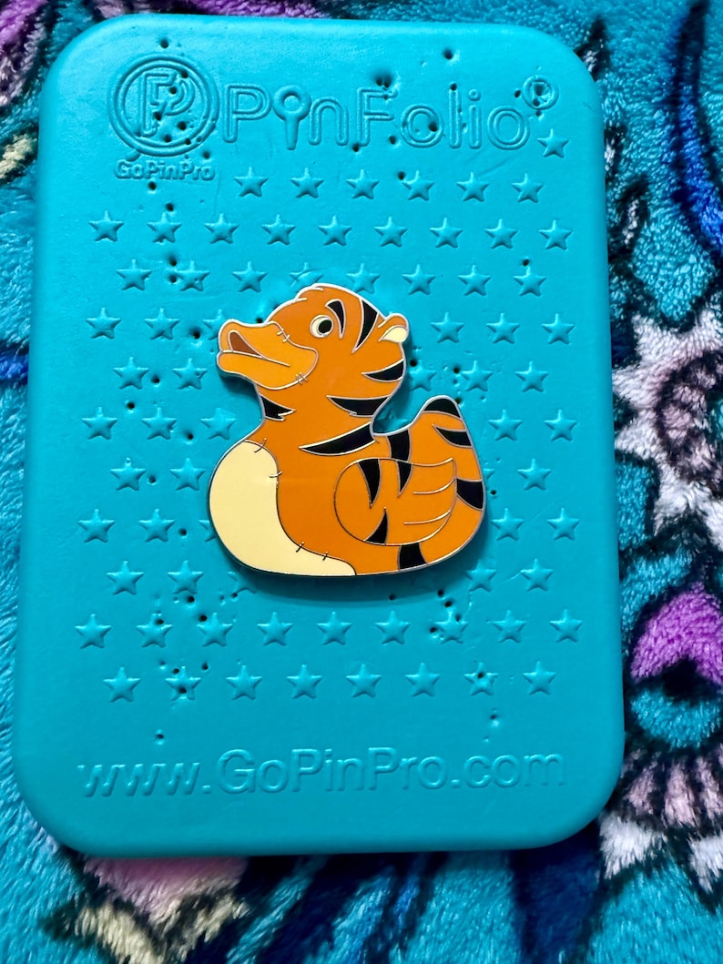 May include: A pin featuring a cartoon tiger duck design, with orange and black stripes, set against a teal background with star-shaped cutouts. The pin is on a teal PinFolio case. The website www.GoPinPro.com is visible.