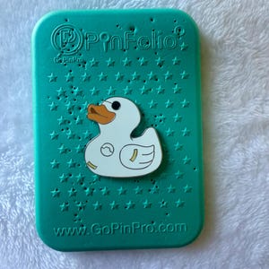 May include: A teal PinFolio pin display case with a white duck pin. The case has a star pattern and the text "PinFolio" and "www.GoPinPro.com". The duck pin has an orange beak and a small design on its body.