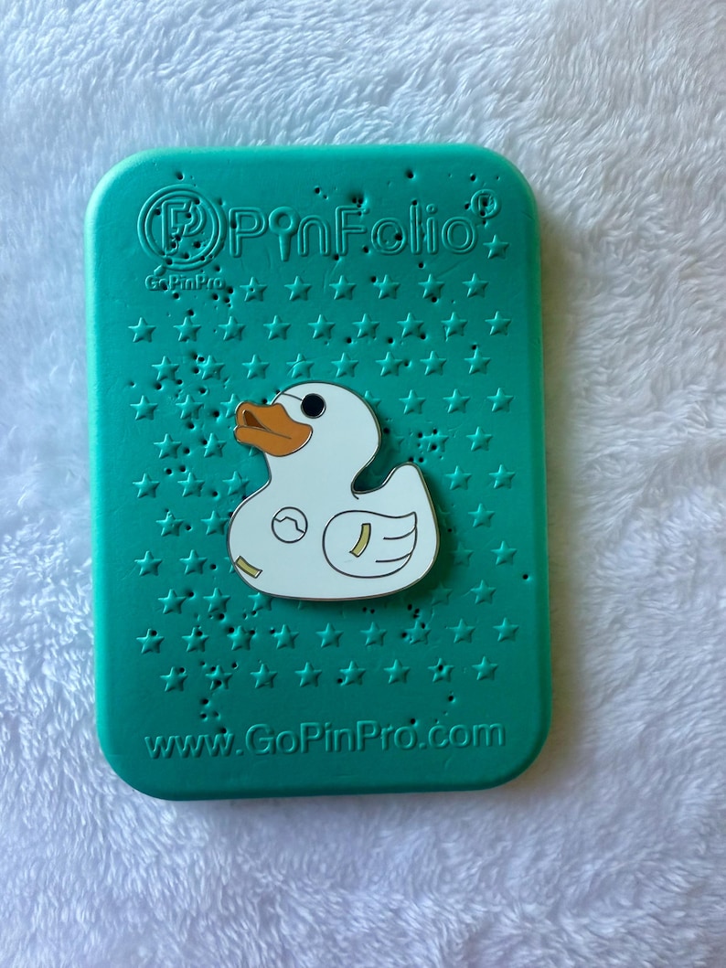 May include: A teal PinFolio pin display case with a white duck pin. The case has a star pattern and the text "PinFolio" and "www.GoPinPro.com". The duck pin has an orange beak and a small design on its body.