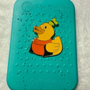 May include: A teal PinFolio pin display case with a yellow duck pin. The duck pin has a red scarf and a green hat. The case has star-shaped holes and the text "www.GoPinPro.com".