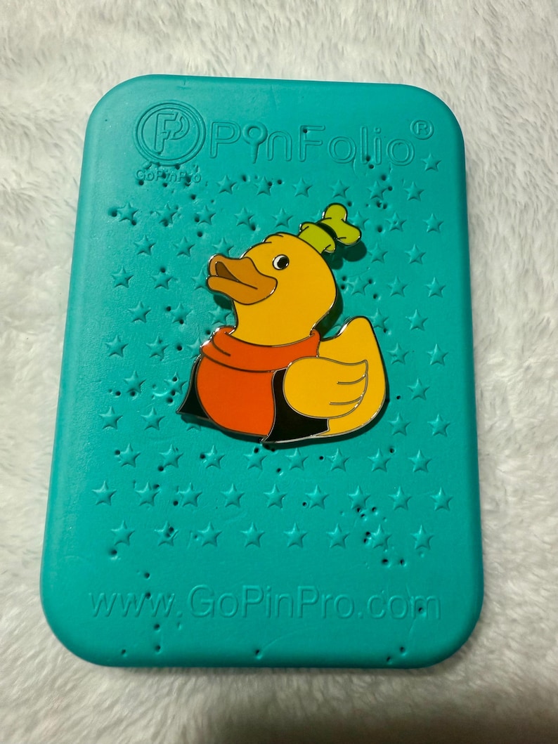 May include: A teal PinFolio pin display case with a yellow duck pin. The duck pin has a red scarf and a green hat. The case has star-shaped holes and the text "www.GoPinPro.com".