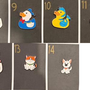 May include: A collection of colorful cartoon animal-shaped pins. The pins include a white duck, a duck wearing a blue sailor outfit, a yellow duck with blue hair, a pink duck with white stripes, a duck with a pink bow, a cat, and a dog.