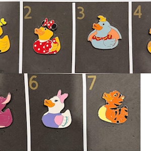 May include: Collection of seven cartoon duck pins. Each pin features a different character design, including a yellow duck with an orange shirt, a duck with a red polka dot dress, and a blue elephant duck. The pins are on a black background.