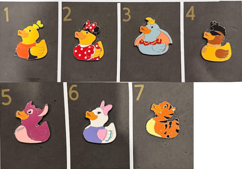 May include: Collection of seven cartoon duck pins. Each pin features a different character design, including a yellow duck with an orange shirt, a duck with a red polka dot dress, and a blue elephant duck. The pins are on a black background.