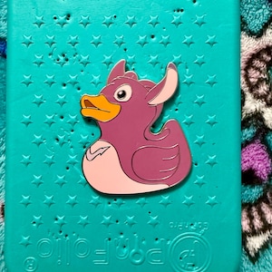 May include: A purple duck pin with a yellow beak and white accents is centered on a teal background covered in star shapes. The pin is a cartoon-style design. The background has the text "www.GoPro.com".