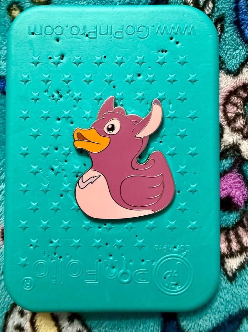 May include: A purple duck pin with a yellow beak and white accents is centered on a teal background covered in star shapes. The pin is a cartoon-style design. The background has the text "www.GoPro.com".