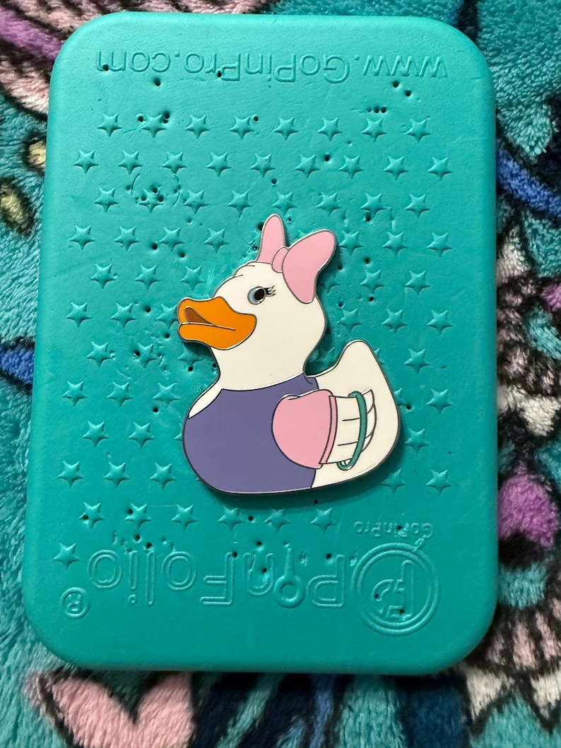 May include: A teal-colored case with a star pattern and a cartoon duck pin. The duck pin has a white body, orange beak, and wears a pink bow, purple top, and pink heart-shaped accessory. The case has the text "www.Gopinpro.com".
