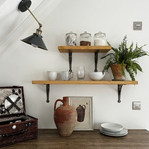 May include: A white wall with two wooden shelves with black metal brackets. The shelves are decorated with glass jars, a fern plant, a ceramic pitcher, a framed print, and white dishes. A brown wooden countertop with a black corner and two black stools is in the foreground.