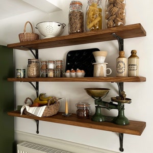 May include: Three wooden shelves with various kitchen items, including jars of pasta, cereal, and spices, a white colander, a black baking pan, a white ceramic mug, a green scale, and a wicker basket with bananas.