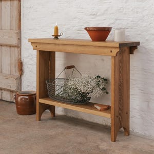 Könnte beinhalten: Ein hellbrauner Holztisch mit einem unteren Regal, das einen Drahtkorb mit weißen Blumen enthält. Eine Terrakotta-Schüssel, ein weißer Keramiktopf und eine brennende Kerze in einem Messinghalter stehen oben drauf. Ein brauner Keramiktopf steht auf dem Boden.