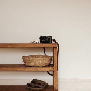 May include: A wooden shelf with two shelves, a woven basket, a pair of brown sandals, and a black camera with a red button on top. The shelf is on wheels and has a white background.