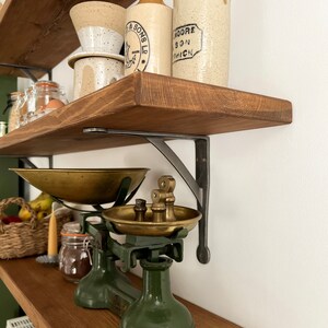 May include: A vintage green kitchen scale with brass bowls and weights sits on a wooden shelf with two antique glass bottles and a ceramic coffee filter. The shelf is supported by black metal brackets.