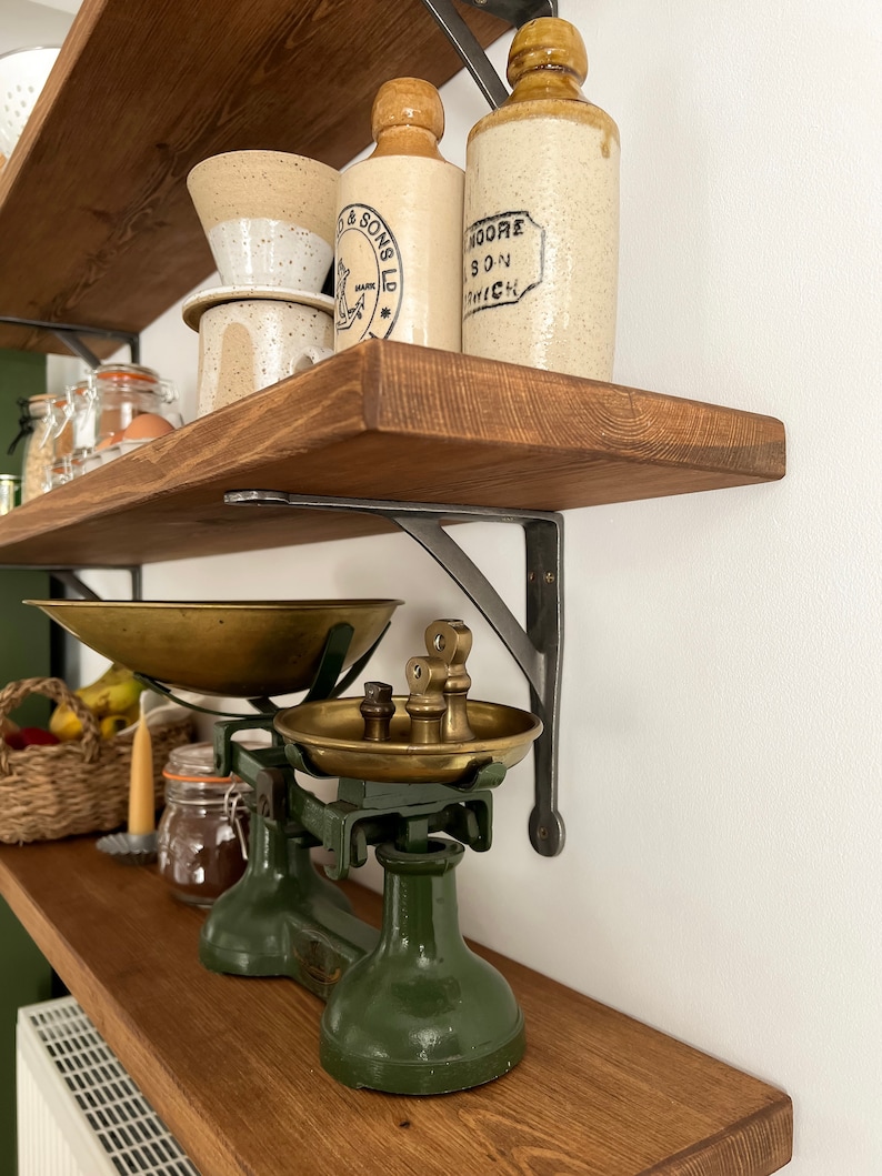 May include: A vintage green kitchen scale with brass bowls and weights sits on a wooden shelf with two antique glass bottles and a ceramic coffee filter. The shelf is supported by black metal brackets.